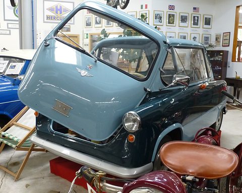 Pale and dark blue small car with its nose door lifted, a motorcycle saddle in the foreground. Opens a larger view