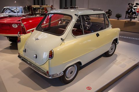 Cream small car from the rear three-quarter on a white plinth, red sports cars and engines behind. Opens a larger view