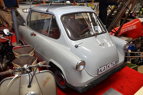 Pale grey small car with plate KA-K 7439 on a red carpet, scooters and a fire engine around it. Opens a larger view