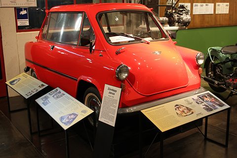 Bright red small car with a front-opening nose door, information boards round it and a motorcycle beside. Opens a larger view