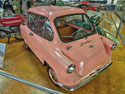 Pink small car with its door toward the camera on a wooden floor beside dressed mannequins. Opens a larger view