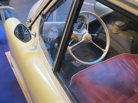 Cream two-spoke steering wheel, simple dashboard and red bench seat seen past the open door frame. Opens a larger view