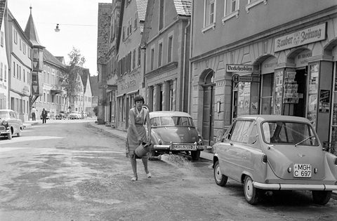 Black-and-white street of old houses, a woman with a watering can, a Janus and a DKW at the kerb.