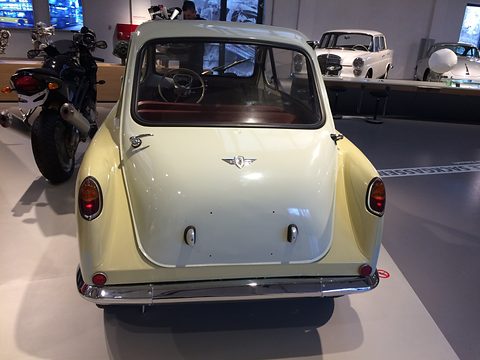 Cream and white small car seen square from behind, its rear door with a winged badge, a motorcycle beside. Opens a larger view