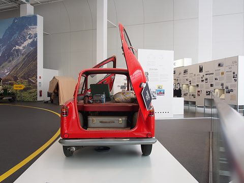 Red small car on a white plinth from behind, its rear door raised over a suitcase and bedding. Opens a larger view