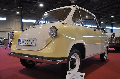 Cream and white small car on a red carpet with a Euroring plate and a show card at its wheel. Opens a larger view
