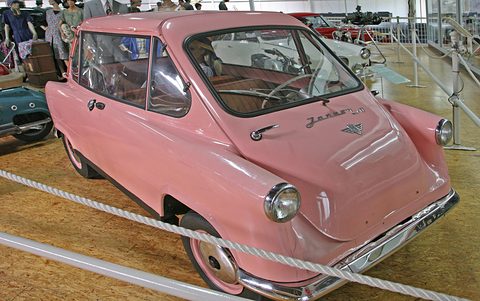 Pink small car behind a rope barrier, its front door and Janus script toward the camera, motorcycles behind. Opens a larger view