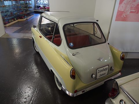 Yellow and white small car from behind on a dark floor, its rear door carrying a “Zündapp Janus” plate. Opens a larger view