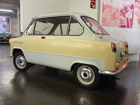 Yellow and white small car in side view on a shiny dark floor, a photograph mural behind. Opens a larger view