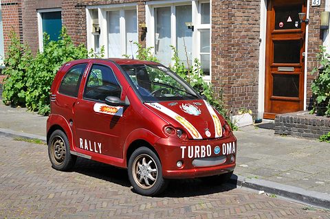 Small red hatchback with white stripes, flame decals and RALLY on its doors, parked on brick paving by a brick house. Opens a larger view