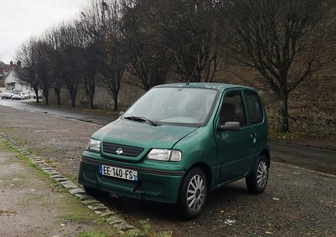 A small green two-door hatchback microcar with an oval grille badge parked on gravel beside a row of trees. Opens a larger view