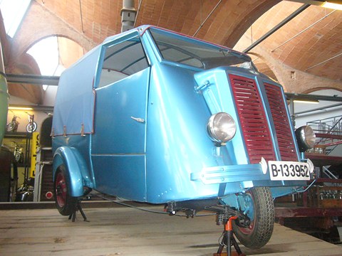 Pale blue three-wheeled van with a canvas-covered rear, red louvred grille and single front wheel, on a stand under brick vaults. Opens a larger view