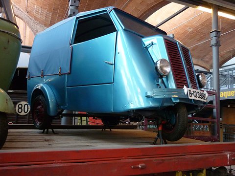 Pale blue three-wheeled van with a canvas rear body, red grille and chrome headlamp, raised on a museum stand. Opens a larger view