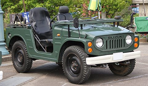 Dark green open small 4x4 with black seats and a roll bar, round headlamps and a white bumper, parked on a car park by trees. Opens a larger view