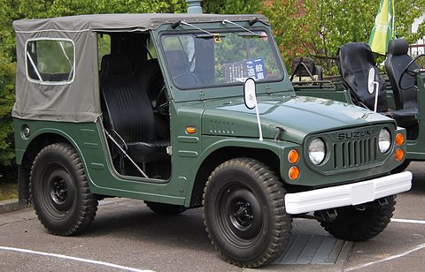 Dark green small 4x4 with a grey canvas top, round headlamps, a slotted grille and a white bumper, on a car park. Opens a larger view