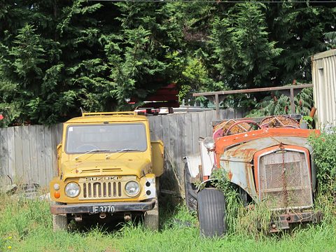 Faded yellow small 4x4 with a slotted grille parked in long grass next to a rusting orange old car body, by a wooden fence and trees. Opens a larger view