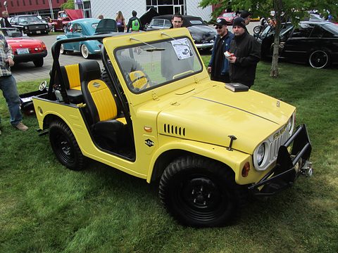 Yellow open small 4x4 with yellow seats, a black roll cage and a front bull bar, on grass with visitors and classic cars behind. Opens a larger view