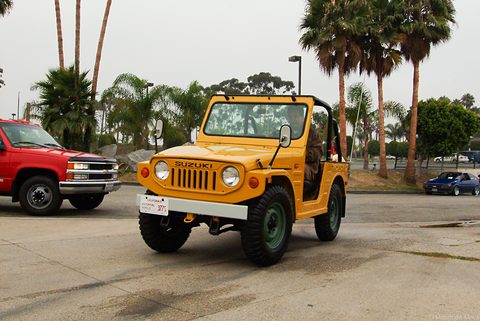 Yellow small 4x4 with green wheels, a roll bar and a slotted grille, parked on a concrete lot with palm trees and a red pick-up behind. Opens a larger view