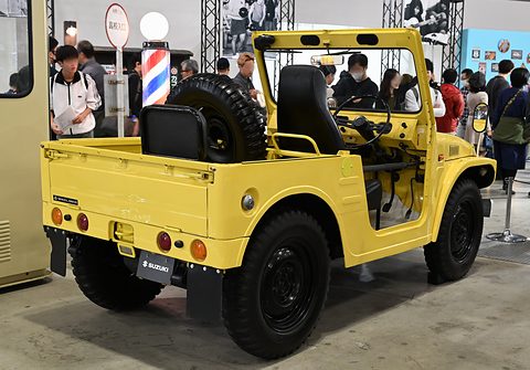 Rear quarter of a yellow open small 4x4 with a spare wheel and black seats in the tub, a barber’s pole and visitors behind. Opens a larger view
