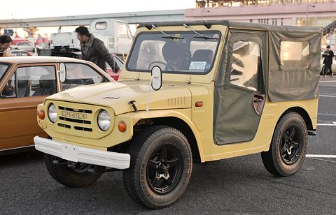 Sand-coloured small 4x4 with a canvas top and canvas doors, round headlamps and black wheels, on a car park among other cars. Opens a larger view