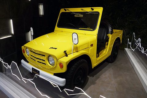 Yellow open small 4x4 with a spare wheel in the tub, round headlamps and a white bumper, lit on a black stand with white line graphics. Opens a larger view