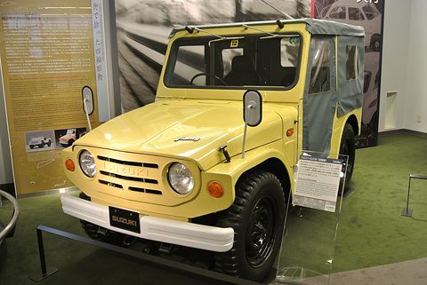 Yellow small 4x4 with a canvas top, round headlamps and a white bumper, on a green museum floor beside an information panel. Opens a larger view