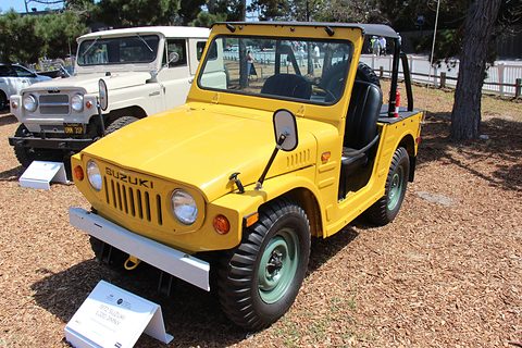 Yellow open small 4x4 with a soft-top frame, round headlamps and a white bumper on bark chippings, a white 4x4 behind. Opens a larger view