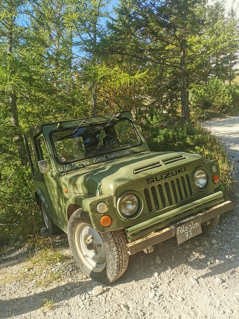 Weathered olive green small 4x4 parked on a gravel mountain track beside trees and bushes.