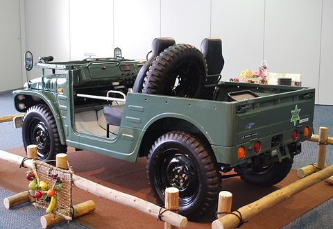 Rear quarter of a green open small 4x4 with a spare wheel standing in the load tub, on a stand with bamboo rails and a basket of fruit. Opens a larger view