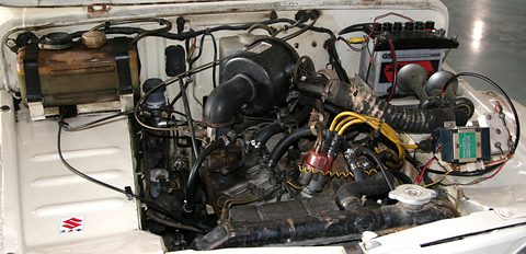 Open bonnet showing a compact engine with a black air intake, yellow ignition leads, a radiator and wiring against white bodywork. Opens a larger view