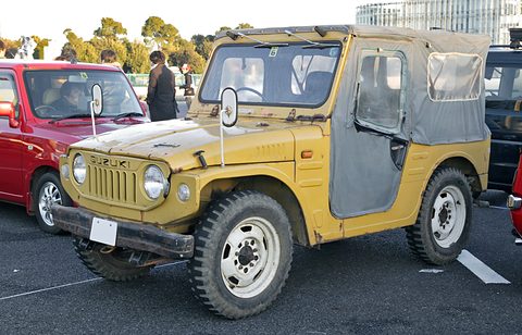 Mustard small 4x4 with a grey canvas top and doors, vertical grille slots and round headlamps, beside a red hatchback on a car park. Opens a larger view