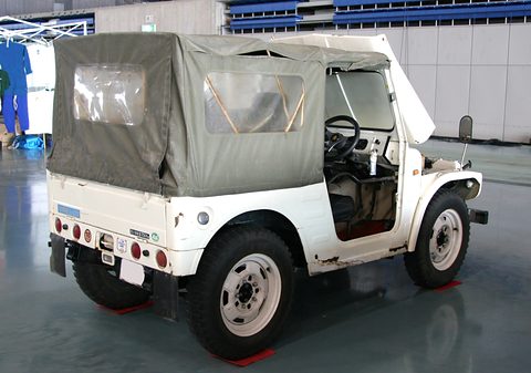 White small 4x4 with a grey canvas top and rear curtain, round red tail lamps and a black bumper, in an indoor hall. Opens a larger view