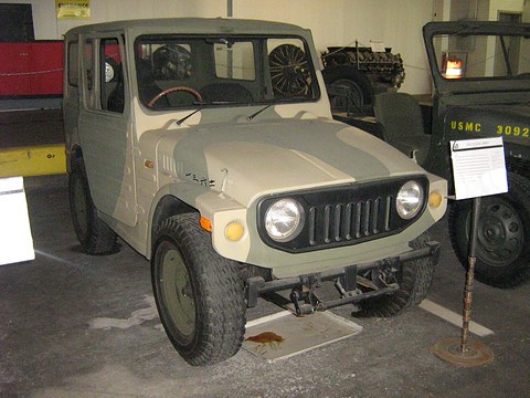 Boxy small 4x4 with a steel roof and camouflage paint, round headlamps and a slotted grille, beside an information stand in a museum. Opens a larger view