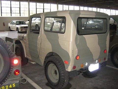 Rear quarter of a boxy small 4x4 with a steel roof, painted in grey and green camouflage, with a spare wheel on the tailgate. Opens a larger view
