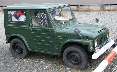 Dark green boxy small 4x4 with a steel roof, round headlamps and a white bumper, parked on gravel by a red and white barrier.