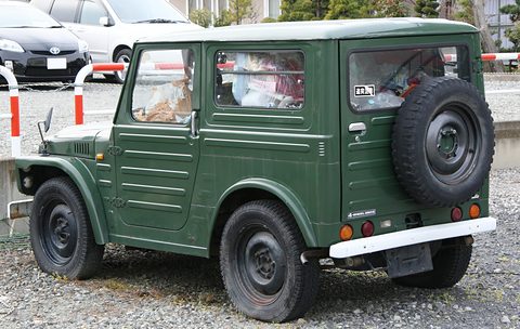 Rear quarter of a dark green boxy small 4x4 with a spare wheel on the tailgate and a white bumper, parked on gravel. Opens a larger view
