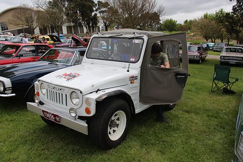 White small 4x4 with a soft top and a half-open canvas door, a man climbing in, parked on grass among classic cars. Opens a larger view