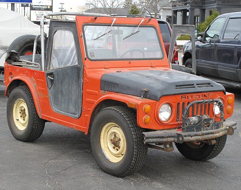 Orange small 4x4 with a black bonnet, cream wheels, steel half-doors, a roll cage and a front winch, parked on tarmac. Opens a larger view