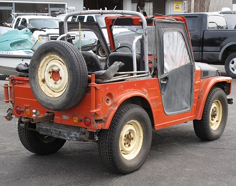 Rear quarter of an orange small 4x4 with a cream spare wheel on the tail, steel half-doors and a roll cage, on tarmac. Opens a larger view