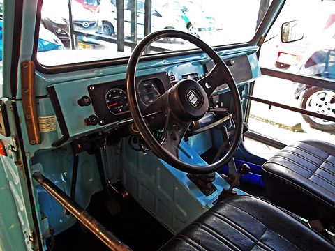 Pale blue painted dashboard with round instruments, a thin black steering wheel and black vinyl seats, seen through the open door. Opens a larger view