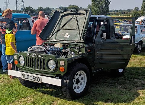 Dark green small 4x4 with its bonnet propped open and doors open, a British number plate, on grass with visitors and cars behind. Opens a larger view