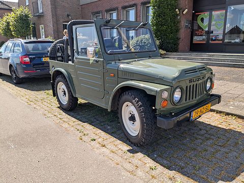 Olive green open small 4x4 with a black roll bar and white wheels, parked on brick paving in front of shops. Opens a larger view
