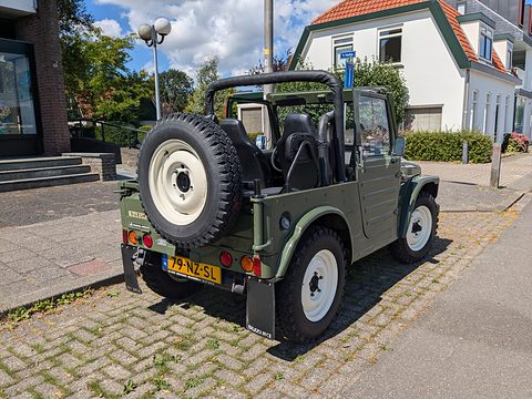 Rear quarter of an olive green open small 4x4 with a white-rimmed spare wheel on the tailgate and a Dutch plate, by a white house. Opens a larger view