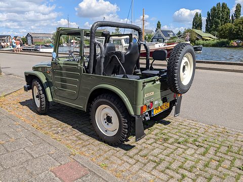 Side view of an olive green open small 4x4 with a roll bar and rear spare wheel, parked on cobbles by a waterway with boats. Opens a larger view