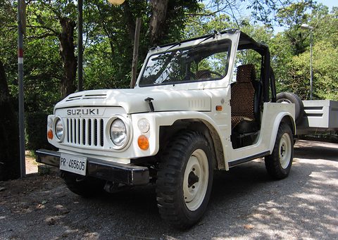 White open-topped small 4x4 with a roll bar, round headlamps and a slotted grille, parked on a gravel lane in the shade of trees. Opens a larger view