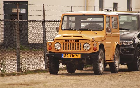 Mustard-yellow boxy small 4x4 with a steel roof, round headlamps and a slotted grille, parked on a gravel yard by a wire fence. Opens a larger view