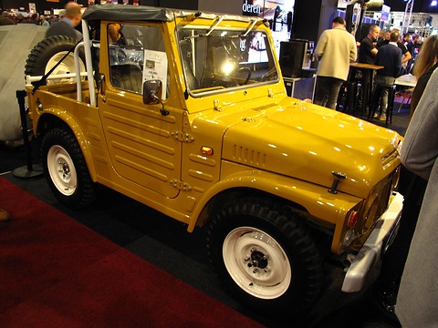 Yellow small 4x4 with a black soft top frame, white wheels and a slotted grille on a show stand, visitors behind. Opens a larger view
