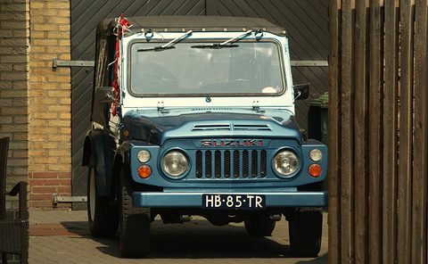 Front view of a blue small 4x4 with a white roof, round headlamps, a slotted grille and a Dutch plate, between a brick wall and a wooden gate. Opens a larger view