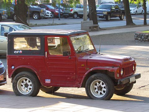 Dark red boxy small 4x4 with a black steel roof and alloy wheels, parked by a pavement with cars and trees behind. Opens a larger view