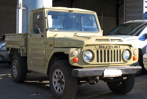 Beige small 4x4 pick-up with a short cab, slotted grille, round headlamps and a flat load bed, parked in a shaded yard. Opens a larger view